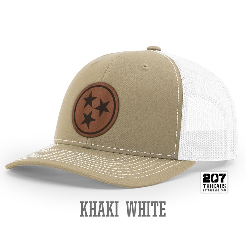 May include: Khaki and white trucker hat with a brown leather patch featuring three stars. The patch is a circle with a stitched edge. The text "207 Threads" is on a white tag attached to the hat.