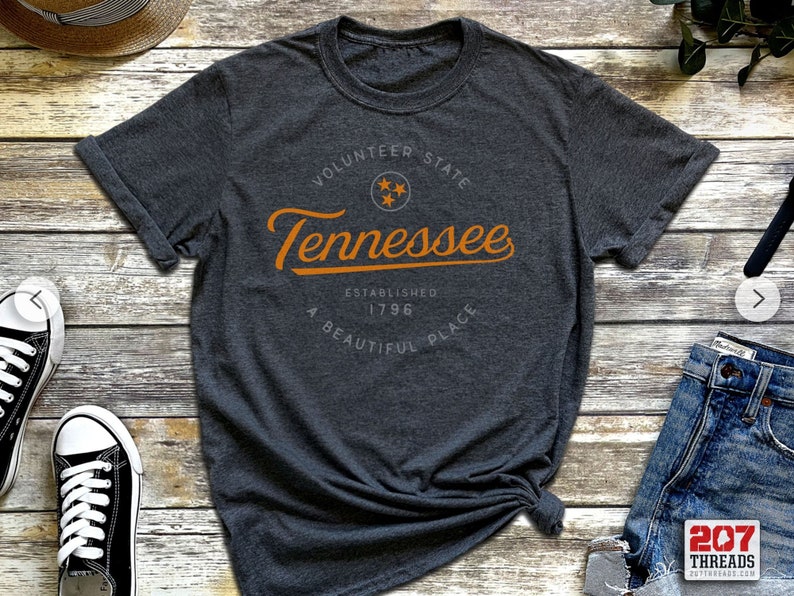 May include: A dark gray t-shirt with an orange graphic design that says "Volunteer State Tennessee Established 1796 A Beautiful Place" with a star inside a circle.