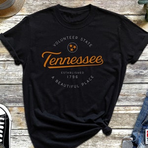 May include: Black t-shirt with a white circle graphic featuring the text "Volunteer State Tennessee Established 1796 A Beautiful Place" in orange text.
