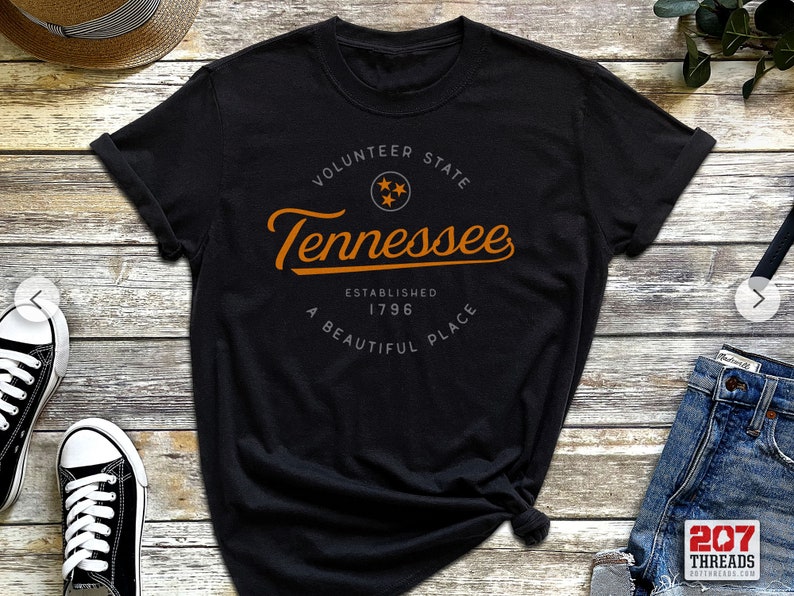 May include: Black t-shirt with a white circle graphic featuring the text "Volunteer State Tennessee Established 1796 A Beautiful Place" in orange text.