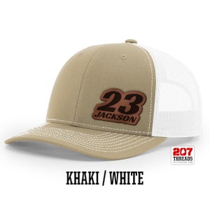 May include: Khaki and white trucker hat with a brown leather patch that says "23 JACKSON" on it.