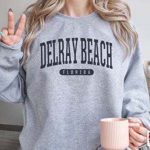 May include: A heather gray sweatshirt with the text "DELRAY BEACH FLORIDA" in black block letters. The sweatshirt is a crew neck style and is being held by a person. A pink mug is in the person's hand.