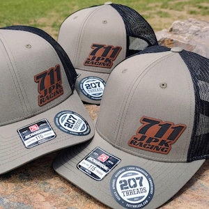 May include: Three khaki trucker hats with black mesh backs. Each hat features a brown leather patch with the text "711 JPK RACING" in black and orange. Additional patches and stickers are visible.