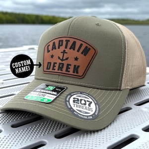 May include: Olive green trucker hat with a brown leather patch. The patch features the text "CAPTAIN DEREK" with an anchor and stars. The hat has a mesh back and a curved brim. The hat also has a "207 Threads" sticker.