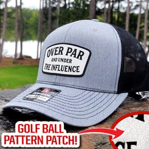 May include: Gray and black trucker hat with the text "OVER PAR AND UNDER THE INFLUENCE" on a white patch. The hat has a golf ball pattern patch and a Richardson 112 Trucker tag.