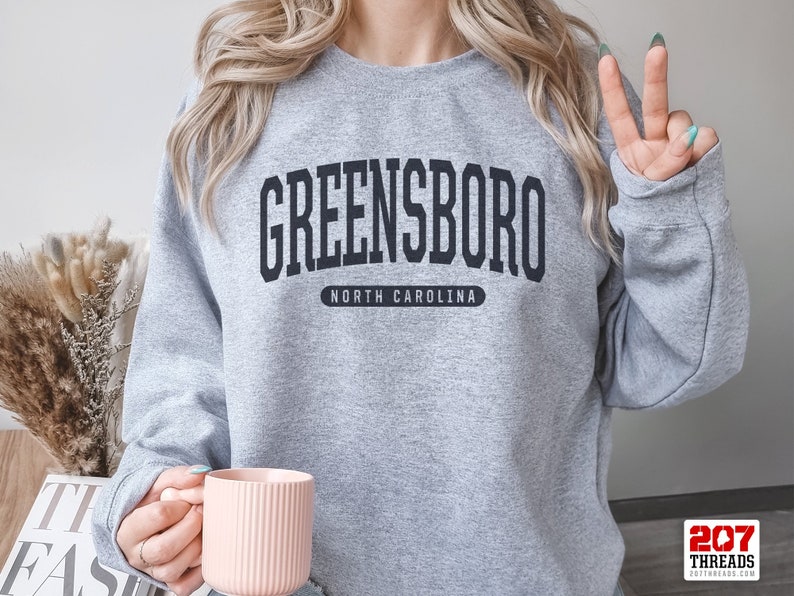 May include: A gray sweatshirt with the word "GREENSBORO" in bold, black letters across the chest, with "NORTH CAROLINA" underneath. The sweatshirt is being worn by a person holding a pink mug.