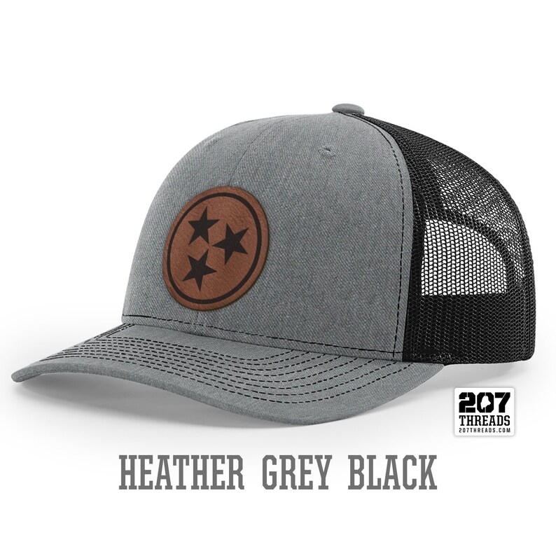 May include: A heather grey and black trucker hat with a brown leather patch featuring three stars. The patch is a circle with a black outline. The hat has a black mesh back. The text "207 Threads" is on a white tag attached to the hat.