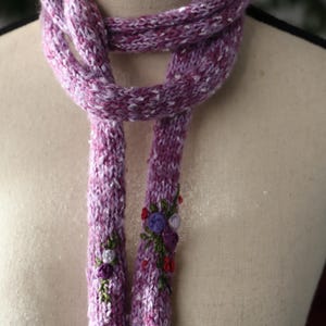 Może przedstawiać: Ręcznie robiony fioletowy szalik z białymi plamkami, ozdobiony haftowanymi detalami kwiatowymi. Szalik jest prezentowany na manekinie, pokazując jego długość i fakturę. Końce szalika zdobią małe, kolorowe kwiatowe zdobienia.