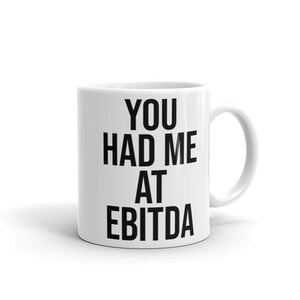 May include: White ceramic coffee mug with black text that reads "You had me at EBITDA".