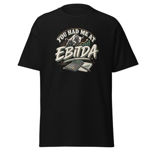 Può includere: Una t-shirt nera a maniche corte con una stampa grafica che riporta il testo "YOU HAD ME AT EBITDA" in bianco e beige. Il design include un toro che carica, un grafico a barre verde in ascesa, una calcolatrice e una penna su un foglio.