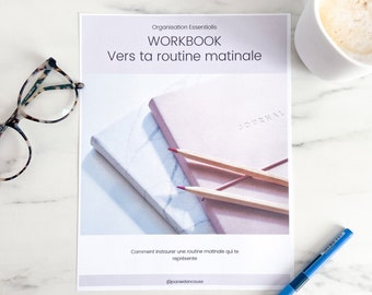 May include: A white workbook with the title "WORKBOOK Vers ta routine matinale" and the text "Comment instaurer une routine matinale qui te représente" and the social media handle "@joaniedancause". The workbook is on a white marble surface with a cup of coffee, a pair of glasses, and a blue pen.
