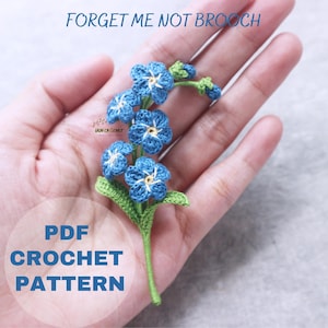 Puede incluir: Un broche de ganchillo de nomeolvides con flores azules y hojas verdes. El texto "FORGET ME NOT BROOCH" está en la parte superior y "PDF CROCHET PATTERN" en la inferior. El broche se sostiene en una mano.