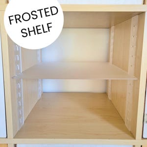May include: A light-colored wooden cabinet interior featuring a horizontal frosted shelf made of semi-transparent material. The shelf is supported by white vertical tracks on both sides. The text FROSTED SHELF is printed in black on a white circle.