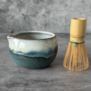 May include: A ceramic matcha bowl with a pour spout, featuring a gradient glaze that transitions from a deep teal at the base to a creamy white at the rim, paired with a light-colored bamboo whisk tied with a thin green thread.