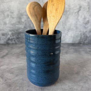 May include: A cylindrical dark blue ceramic utensil holder with a speckled finish and horizontal ribbed texture. Three light-colored wooden kitchen utensils, including two spoons and a flat-edged spatula, are placed inside the container.