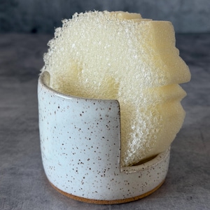May include: A white ceramic sponge holder with brown speckles and a cutout side design, containing a pale yellow, textured, porous cleaning sponge. The holder has a cylindrical shape with a flat base and a smooth, glazed finish on the exterior.
