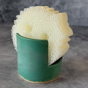 May include: A matte forest green ceramic holder with a U-shaped cutout, containing a stack of three round, pale yellow, textured abrasive sponges. The holder has a smooth, solid-colored exterior and a circular base, designed for upright storage.