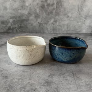 May include: Two ceramic bowls with pouring spouts. One is speckled white, the other is a deep blue with a gold rim. The bowls are round with a small spout on the side. The background is a textured grey.