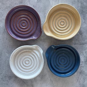 May include: Four ceramic spoon rests in shades of purple, beige, white, and blue. Each rest has a circular design with a small spout. The spoon rests are arranged on a grey surface.