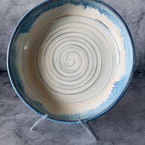 May include: A round ceramic bowl featuring a cream-colored interior with a dark blue spiral pattern at the center. The rim of the bowl is finished with a textured, mottled blue glaze that transitions into the lighter cream base of the vessel.