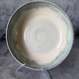 May include: A round ceramic bowl featuring a smooth, cream-colored center with a visible spiral texture. The wide rim is finished with a reactive glaze in shades of sage green and muted teal, creating a soft, blended transition toward the edge.