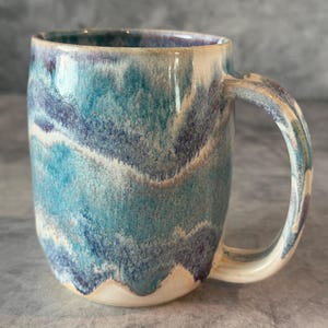 May include: A ceramic mug featuring a reactive glaze with swirling patterns of teal, deep blue, and cream. The mug has a rounded body, a smooth rim, and a curved handle, showcasing a layered, multi-colored finish that transitions from dark to light.
