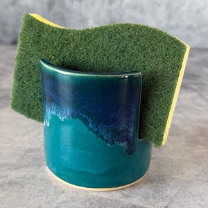 May include: A ceramic sponge holder with a glossy finish featuring a deep blue upper section that transitions into a vibrant teal base. The holder has a curved, open-top design and contains a rectangular green scouring pad with a yellow sponge layer.