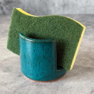 May include: A teal ceramic sponge holder with a speckled finish containing a rectangular yellow and green scouring sponge. The holder has a curved, open-front design with a smooth glazed surface and a visible terracotta-colored base rim.