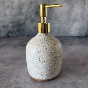 May include: A ceramic soap dispenser with a speckled off-white finish and a shiny gold-toned metal pump mechanism. The vessel has a rounded, bulbous shape with a smooth, glossy texture and a visible brown base rim.