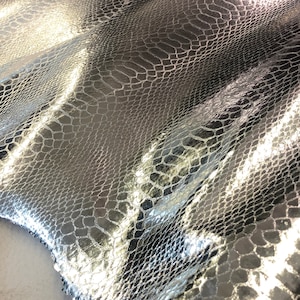 May include: Silver snakeskin fabric with a textured, scaly pattern. The fabric is folded and has a reflective, metallic sheen.