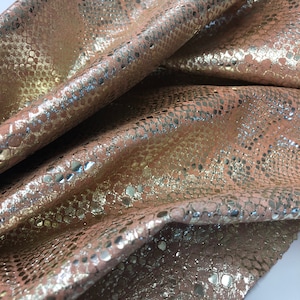 May include: A close-up of a piece of pink snakeskin leather with a metallic gold finish. The leather has a textured surface and a subtle sheen.