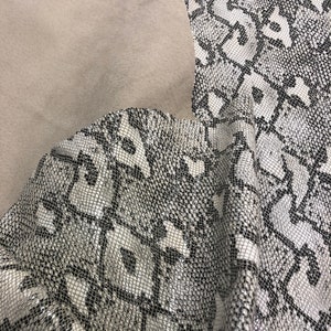 May include: A close-up of a white and black snakeskin fabric with a textured pattern. The fabric is folded and has a smooth, light brown backing.