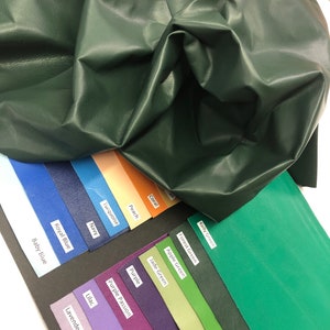 May include: A green leather swatch laid over a color chart with swatches of various colors, including blue, turquoise, peach, coral, kelly green, forest green, apple green, jade green, purple, purple passion, lilac, and lavender.