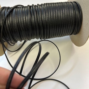 May include: A spool of black leather cord, wound around a cardboard core. The cord is smooth and has a slight sheen.