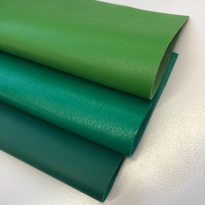 May include: Three sheets of green leather in varying shades are layered on top of each other. The top sheet is a bright green, the middle is a medium green, and the bottom is a deep forest green. The leather has a textured surface.