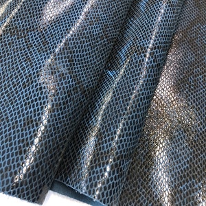 May include: Close-up of a dark blue snakeskin leather fabric with a shiny finish. The fabric is folded in a way that shows the texture and pattern of the leather.