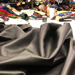 May include: A close-up of a large piece of black leather fabric folded on a white surface. The fabric has a textured surface and is wrinkled. There are various rolls of leather and other materials in the background.