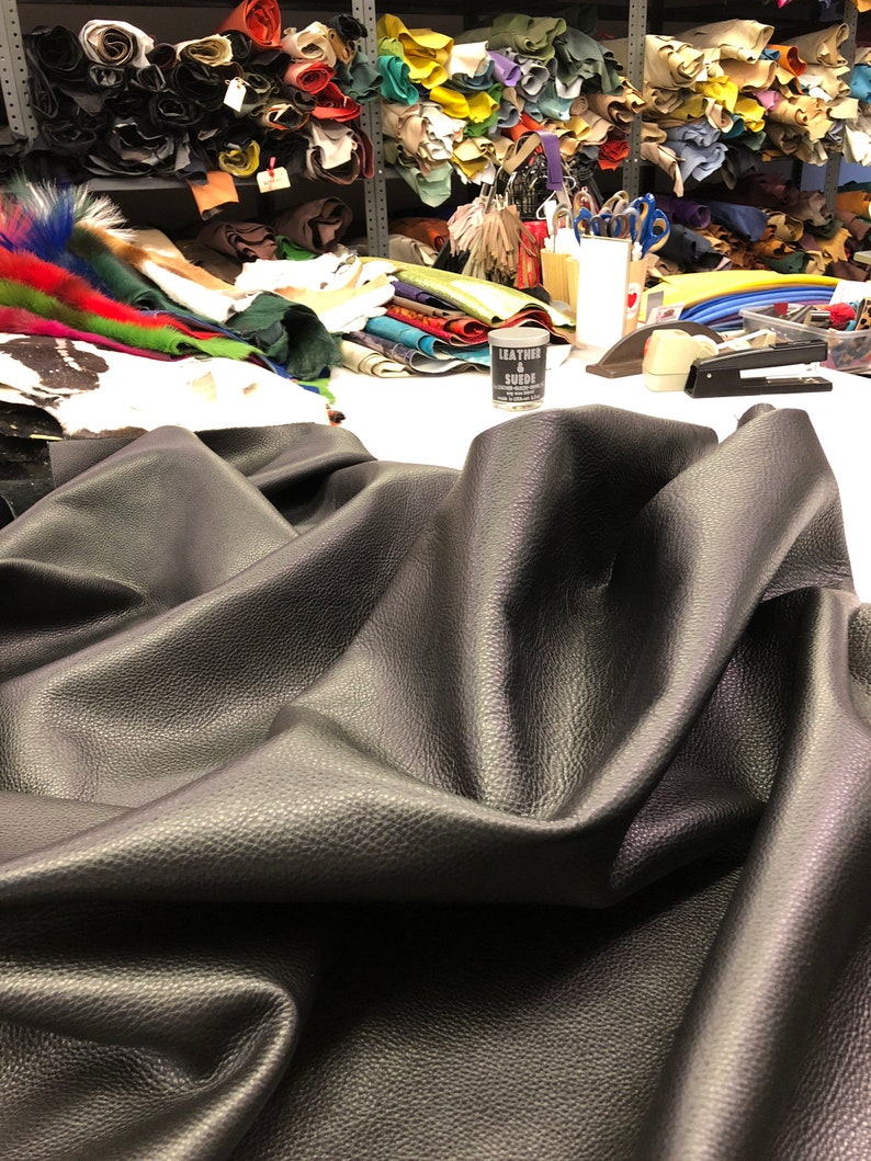 May include: A close-up of a large piece of black leather fabric folded on a white surface. The fabric has a textured surface and is wrinkled. There are various rolls of leather and other materials in the background.