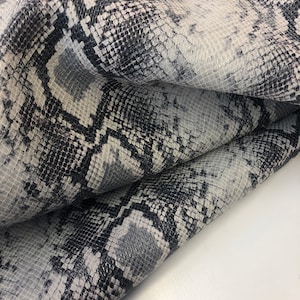 May include: Close-up of a textured snakeskin pattern in shades of black, gray, and white. The material is folded, revealing the intricate scales and color variations. The overall appearance suggests a luxurious or exotic textile.