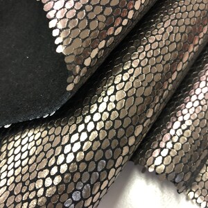 May include: Close-up of a silver snakeskin fabric with a textured pattern. The fabric is folded over itself, showing the underside of the material.