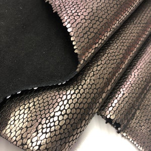 May include: Close-up of a metallic silver snakeskin fabric with a black backing. The fabric has a textured, scaly pattern.