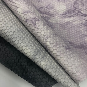 May include: Three pieces of fabric with a textured, scale-like pattern. The top piece is a light purple with a marbled effect. The middle piece is a light gray with a subtle marbled effect. The bottom piece is black.