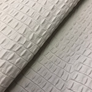 May include: White faux crocodile embossed leather fabric. The fabric has a textured, alligator-like pattern.