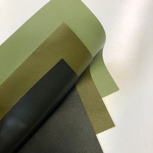 May include: Three pieces of leather in different shades of green and black. The leather is folded in a way that shows the different colors and textures.