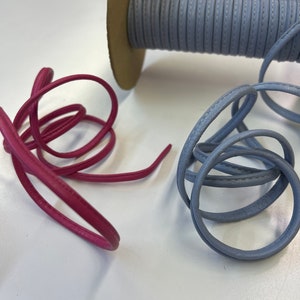 May include: Two spools of leather cord, one in a light blue color and one in a deep pink color. The light blue cord is wound around a cardboard spool. The pink cord is loose and coiled.