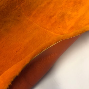 May include: A close-up of a piece of orange fabric with a fuzzy texture on one side and a smooth, brown leather texture on the other side.