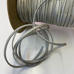 May include: A spool of silver-colored leather cord with a stitched edge. The cord is wound around a cardboard spool.