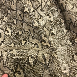 May include: Close-up of a beige and black snakeskin fabric with a geometric pattern. The fabric is textured and has a slight sheen.