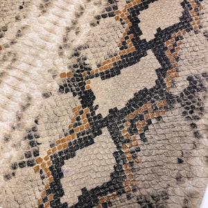 K&ouml;nnte beinhalten: Nahaufnahme eines beigefarbenen Schlangenlederstoffs mit einem schwarzen und braunen Muster. Der Stoff hat eine strukturierte Oberfl&auml;che und ein sich wiederholendes Muster aus Diamanten.