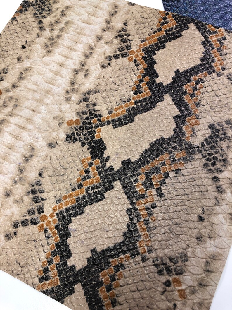 K&ouml;nnte beinhalten: Nahaufnahme eines beigefarbenen Schlangenlederstoffs mit einem schwarzen und braunen Muster. Der Stoff hat eine strukturierte Oberfl&auml;che und ein sich wiederholendes Muster aus Diamanten.